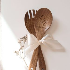 Salabesteck bestehend aus zwei Besteckteilen in dunklem Holz mit Gravur Initalen und Datum und weißer Schleife in einem neutralen Setting