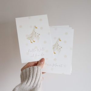 Meilensteinkarten mit dem motiv einer Gans und pastellfarbenen Punkten und dem Schriftzug "hallo Welt ich bin da" von einer Hand haltend vor einem neutralen Hintergrund