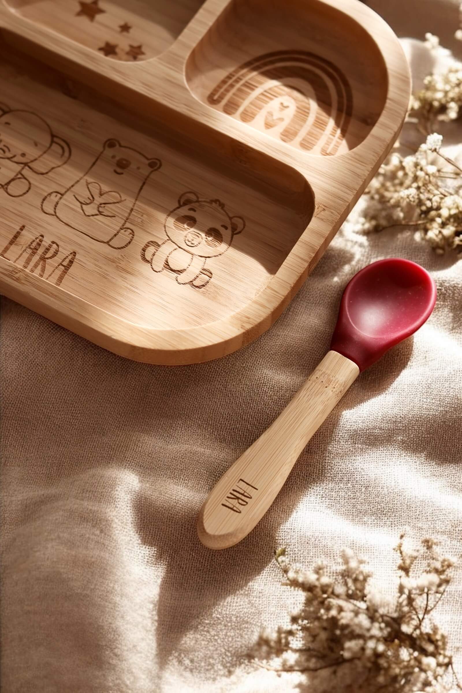 Quadratischer Bambusteller für Babys mit drei Fächern. In den drei Fächern befinden sich Gravuren von Sternen, Regenbogen und Elefant, Bär und Pandabär mit einem Namen darunter. Der Teller steht auf einem Holzbrett mit natürlichem Sonnenlicht.