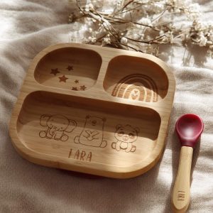 Quadratischer Bambusteller für Babys mit drei Fächern. In den drei Fächern befinden sich Gravuren von Sternen, Regenbogen und Elefant, Bär und Pandabär mit einem Namen darunter. Der Teller steht auf einem Holzbrett mit natürlichem Sonnenlicht.