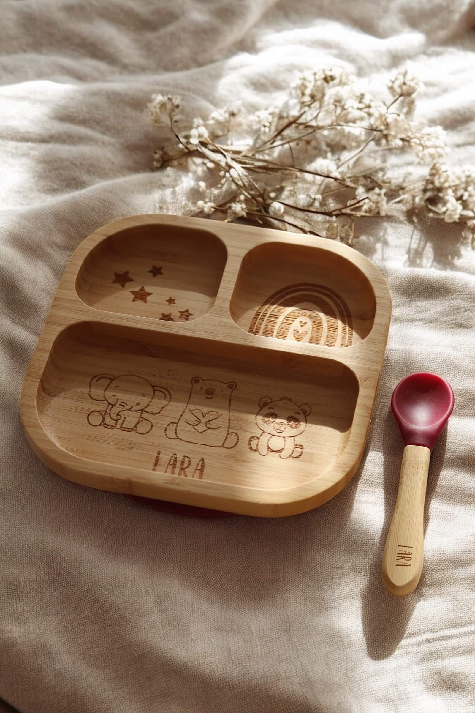 Quadratischer Bambusteller für Babys mit drei Fächern. In den drei Fächern befinden sich Gravuren von Sternen, Regenbogen und Elefant, Bär und Pandabär mit einem Namen darunter. Der Teller steht auf einem Holzbrett mit natürlichem Sonnenlicht.