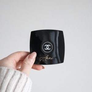schwarzer quadratisches Spiegelgehäuse mit dem Chanel Logo und dem Namen "Anni" in Gold von einer Hand gehalten vor einem neutralen Hintergrund