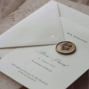 Weißer Papierumschlag auf Stein gelegt mit goldenen Buchstaben auf der Lasche. Die Buchstaben sind die Initialien des Brautpaares und werden durch Heißfolienprägung in das Papier gestanzt.
