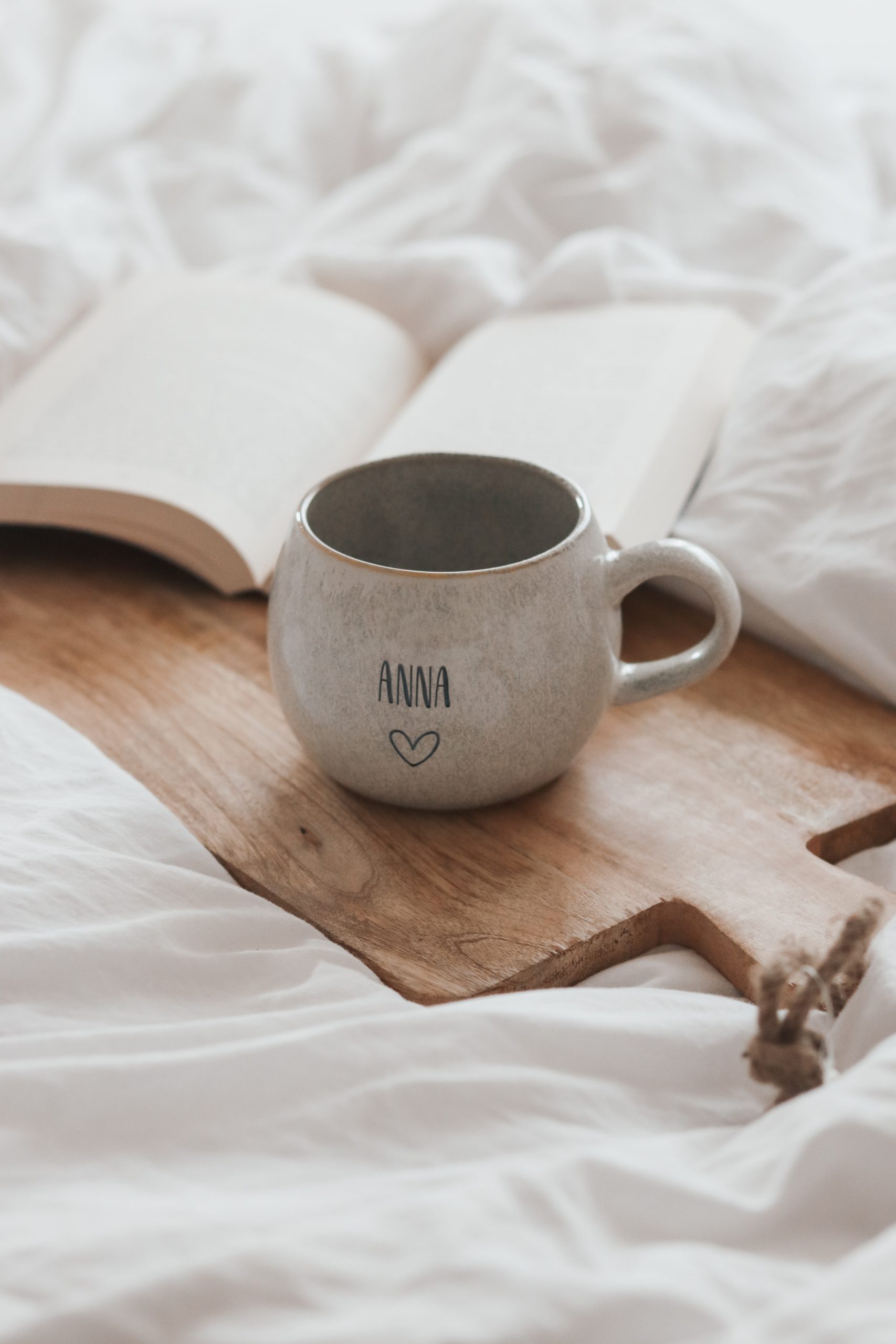 Tasse personalisiert. große beige graue Keramiktasse mit graviertem Wunschtext auf einer weißen Decke mit einem Schneidebrett aus Holz und einem aufgeschlagenen Buch im Hintergrund