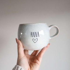 Große runde Tasse aus beigem Keramik gefertigt und der Gravur "Anna" und "Herz" in einer Hand haltend
