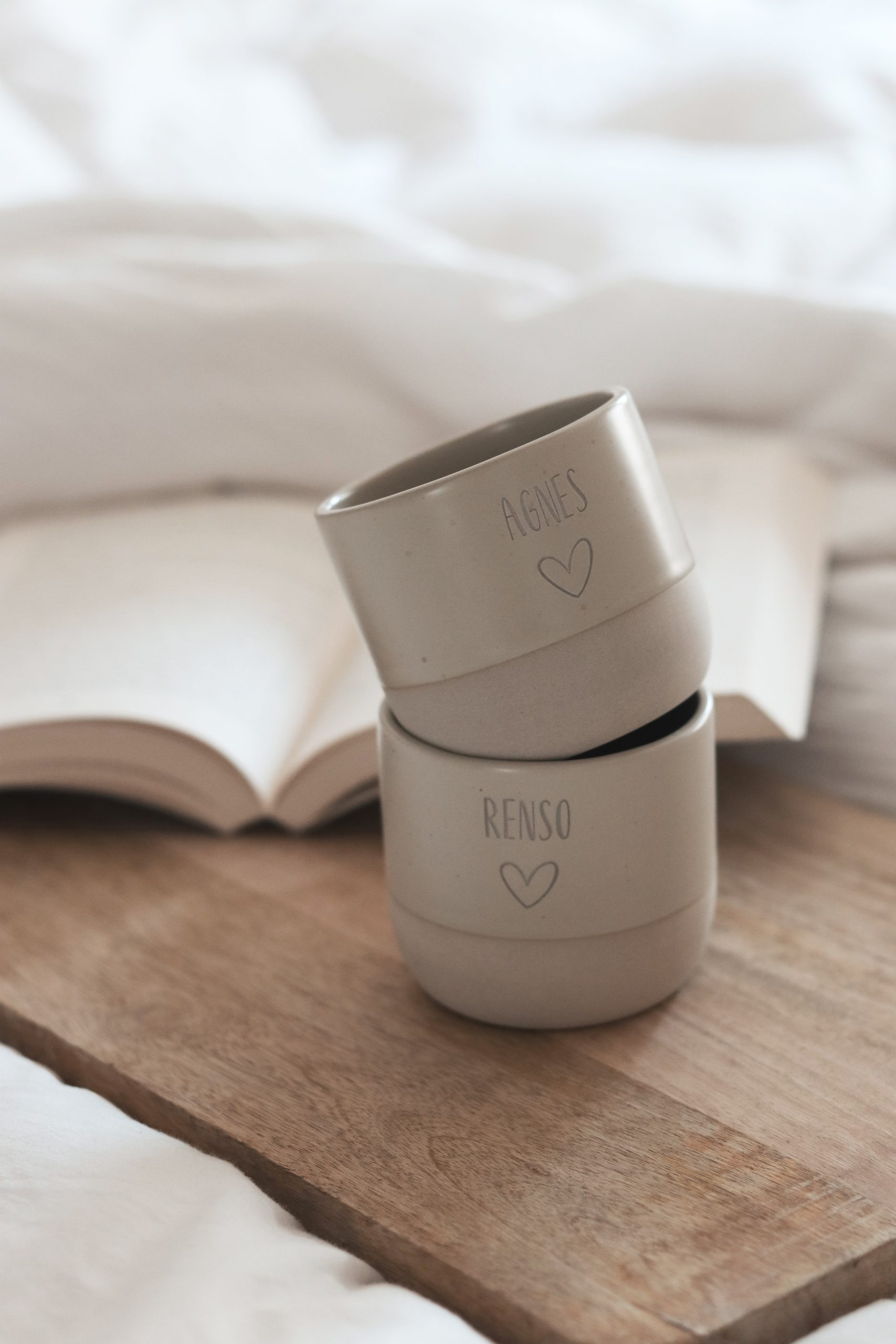 Beige runde Steinguttasse mit Wunschtext und Herz personalisiert darunter ein Savierbrett aus Holz und ein aufgeschlagenes Buch im Hintergrund