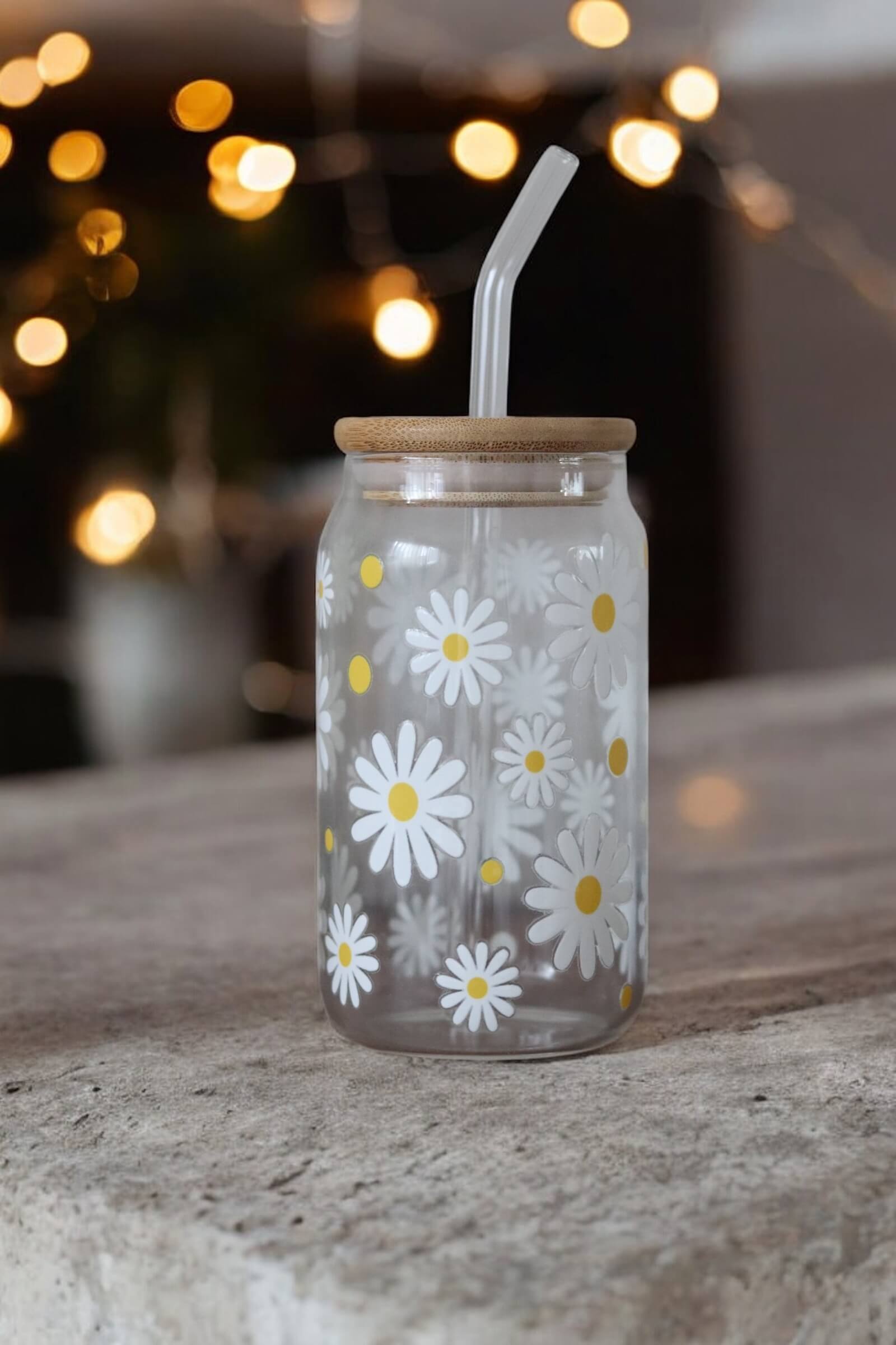 Glas mit Bambusdeckel und Glasstrohhalm und Muster mit weißen Blumen auf einer Betonarbeitsplatte und einer Lichterkette im Hintergrund