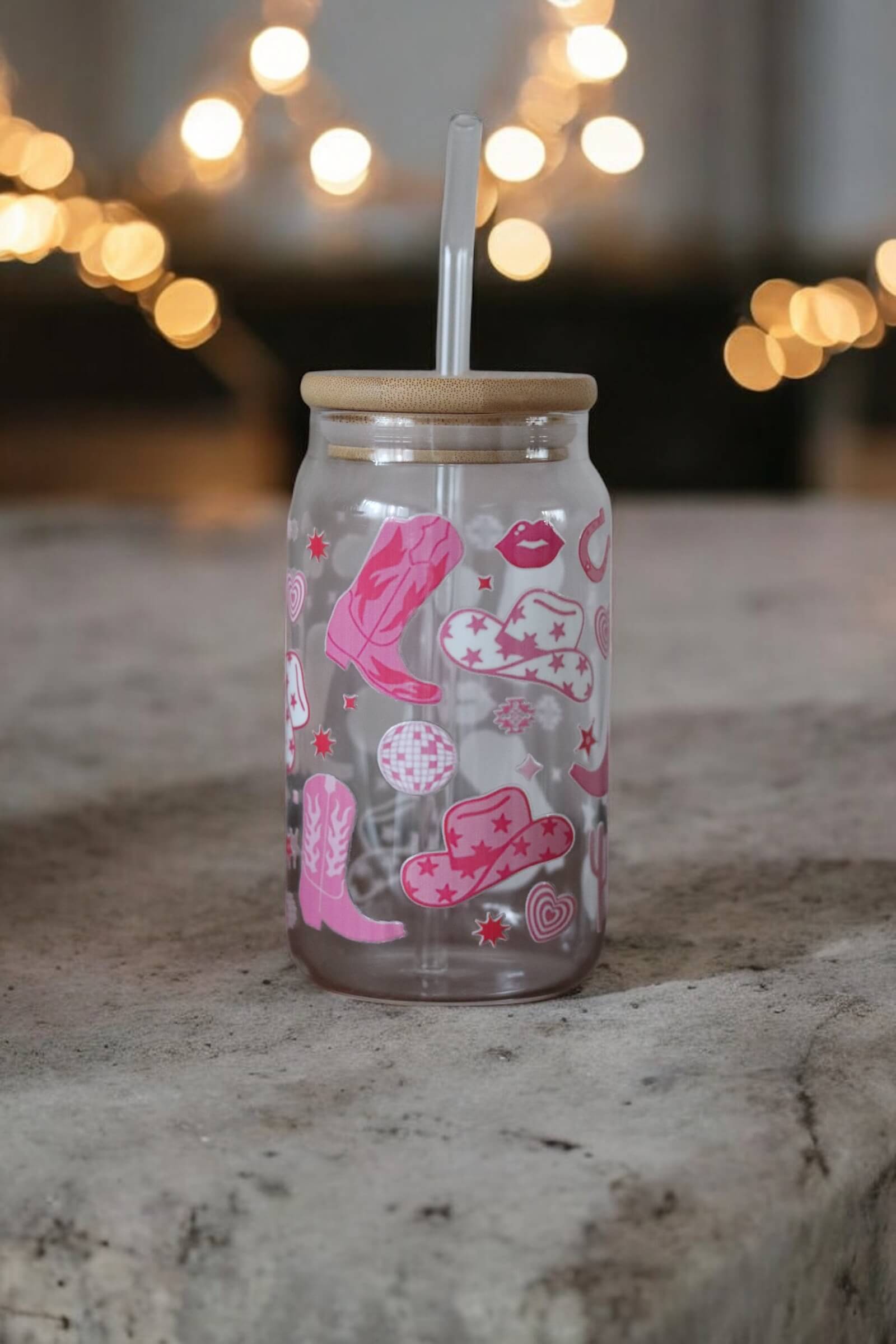 Glas mit Bambusdeckel und Glasstrohhalm und Muster mit pinken Cowboystiefeln und Hüten auf einer Betonarbeitsplatte und einer Lichterkette im Hintergrund