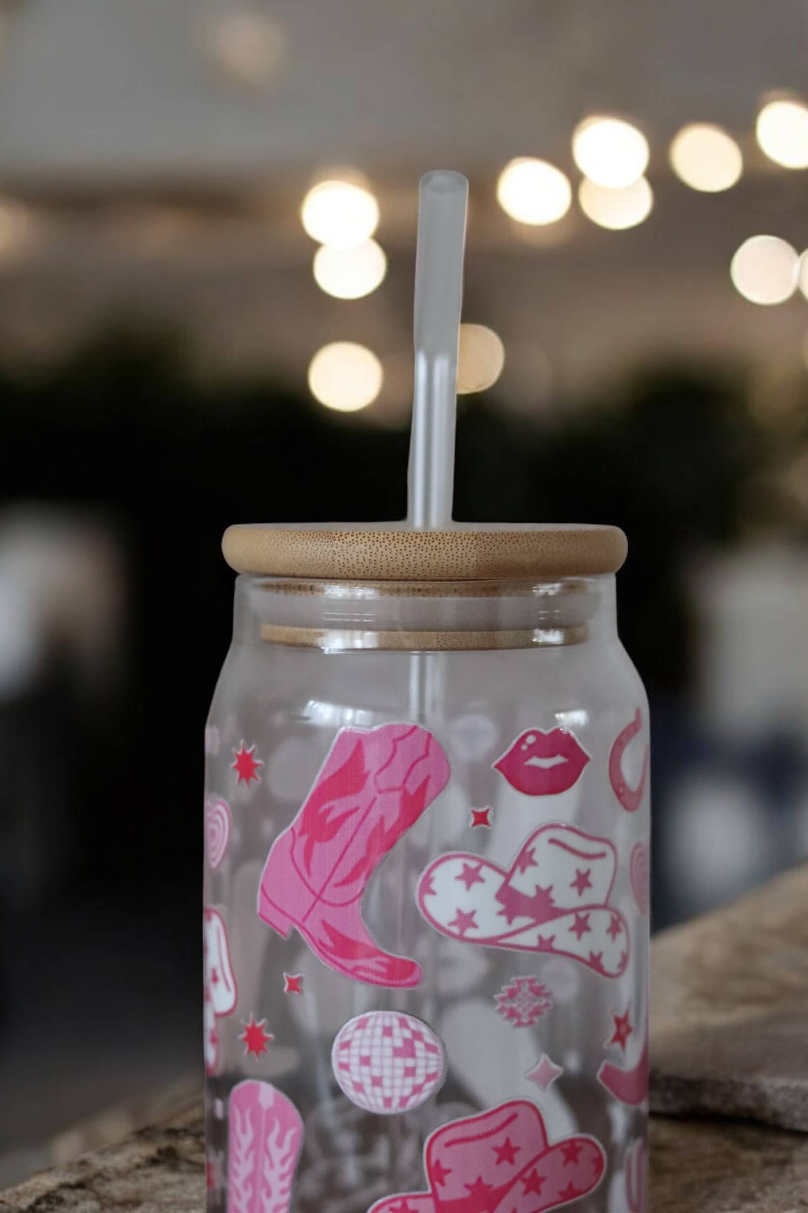Glas mit Bambusdeckel und Glasstrohhalm und Muster mit pinken Cowboystiefeln und Hüten auf einer Betonarbeitsplatte und einer Lichterkette im Hintergrund