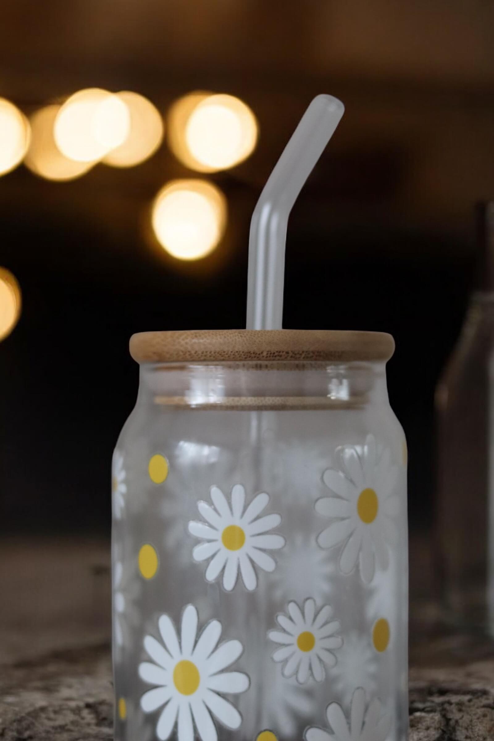 Glas mit Bambusdeckel und Glasstrohhalm und Muster mit weißen Blumen auf einer Betonarbeitsplatte und einer Lichterkette im Hintergrund