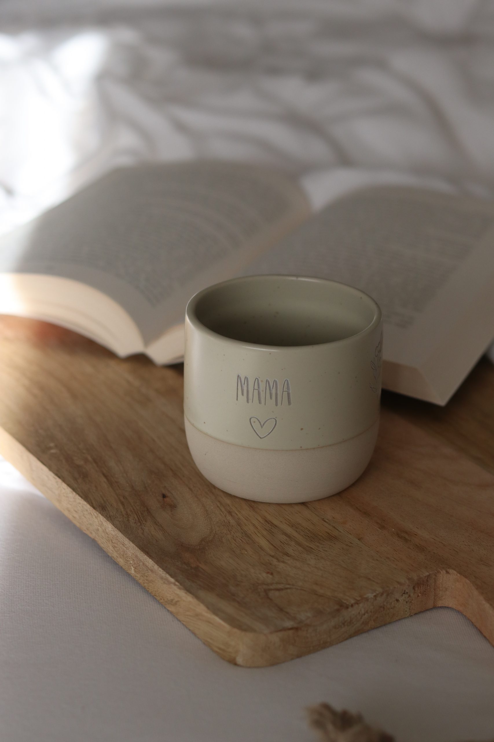 Beige runde Steinguttasse mit Wunschtext und Herz personalisiert darunter ein Savierbrett aus Holz und ein aufgeschlagenes Buch im Hintergrund