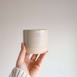 Beige runde Tasse ohne Griff aus Keramik und mit der Gravur "Anna" und dem Symbol "Herz" von einer Hand haltend