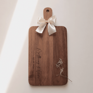 Servierbrett aus Akazienholz mit Griff und personalisierter Gravur und weißer Schleife am Griff in einem neutralen Hintergrund