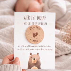 Karte mit einer Entscheidungsmünze aus Holz mit der Gravur "wer ist dran? Mama" im Hintergrund ein schlafendes Baby