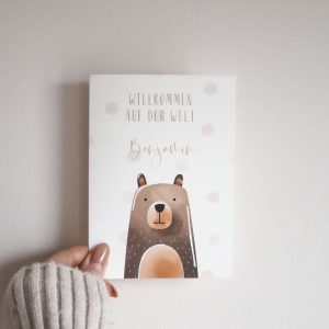 weiße Karte im Postkartenformat mit Bärenmotiv und dem Text "Willkommen auf der Welt Benjamin" von einer Hand gehalten vor einem neutralen Hintergrund