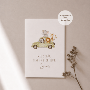 Eine weiße Grußkarte liegt mittig und zeigt eine kleine Illustration eines Autos mit Tieren. Darunter steht der Schriftzug „Wie schön, dass es dich gibt, Lukas“. Rechts neben der Karte liegt ein feiner, getrockneter Zweig, während weiches Licht und sanfte Schatten für eine ruhige, natürliche Atmosphäre sorgen.