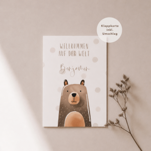 Eine weiße Grußkarte liegt mittig und zeigt eine Illustration aus Aquarell mit einem Bär mit dem dem Schriftzug "Willkommen auf der Welt Benjamin“. Rechts neben der Karte liegt ein feiner, getrockneter Zweig, während weiches Licht und sanfte Schatten für eine ruhige, natürliche Atmosphäre sorgen.