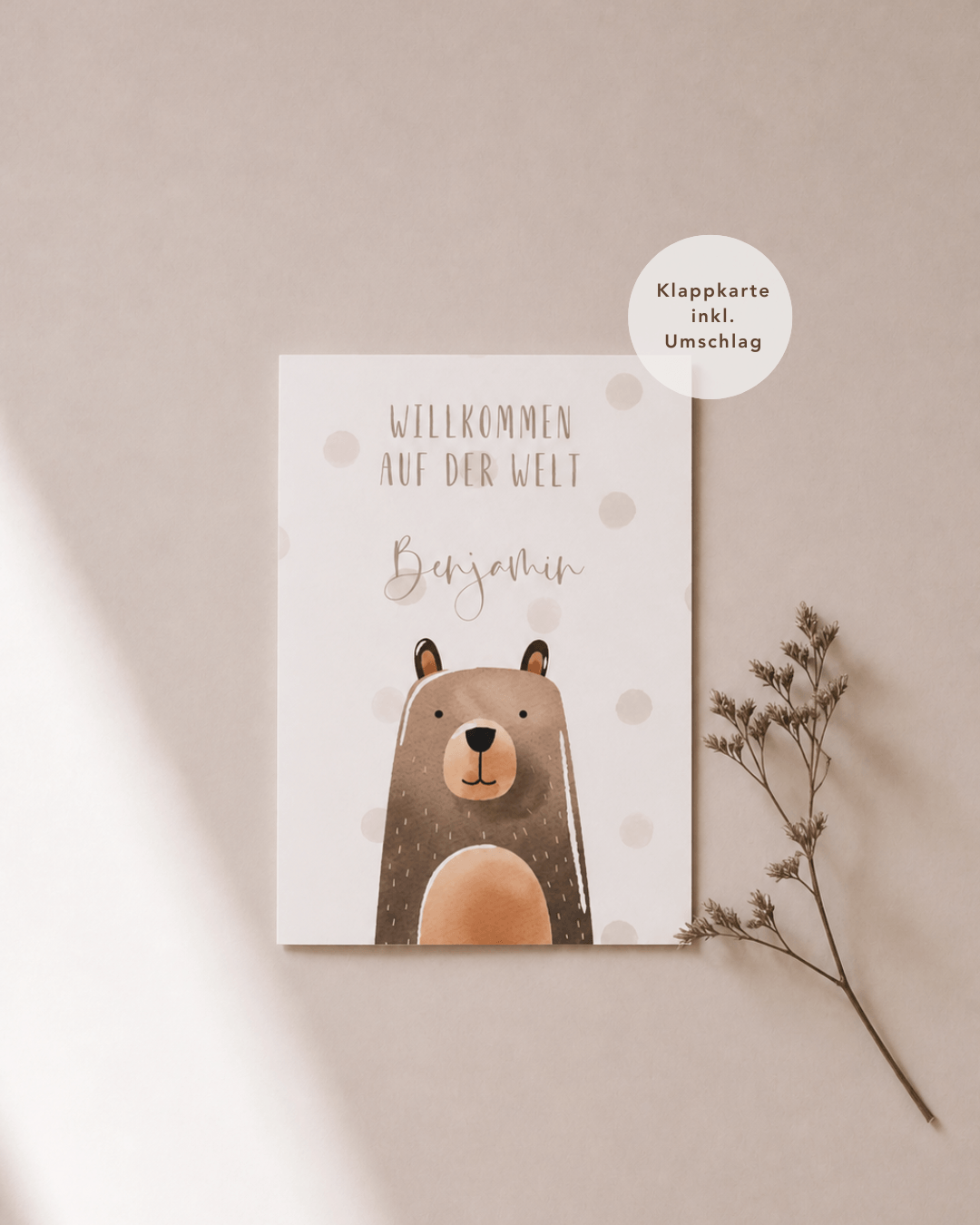 Eine weiße Grußkarte liegt mittig und zeigt eine Illustration aus Aquarell mit einem Bär mit dem dem Schriftzug "Willkommen auf der Welt Benjamin“. Rechts neben der Karte liegt ein feiner, getrockneter Zweig, während weiches Licht und sanfte Schatten für eine ruhige, natürliche Atmosphäre sorgen.