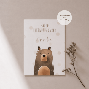 Eine weiße Grußkarte liegt mittig und zeigt eine Illustration aus Aquarell mit einem Bär mit dem dem Schriftzug "Hallo kleines Wunder Annika“. Rechts neben der Karte liegt ein feiner, getrockneter Zweig, während weiches Licht und sanfte Schatten für eine ruhige, natürliche Atmosphäre sorgen.
