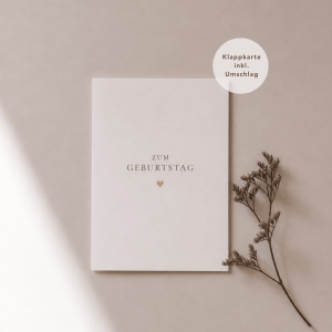 Eine weiße Grußkarte liegt mittig und zeigt den Schriftzug "zum Geburtstag“ mit einem goldenen Herz darunter. Rechts neben der Karte liegt ein feiner, getrockneter Zweig, während weiches Licht und sanfte Schatten für eine ruhige, natürliche Atmosphäre sorgen.