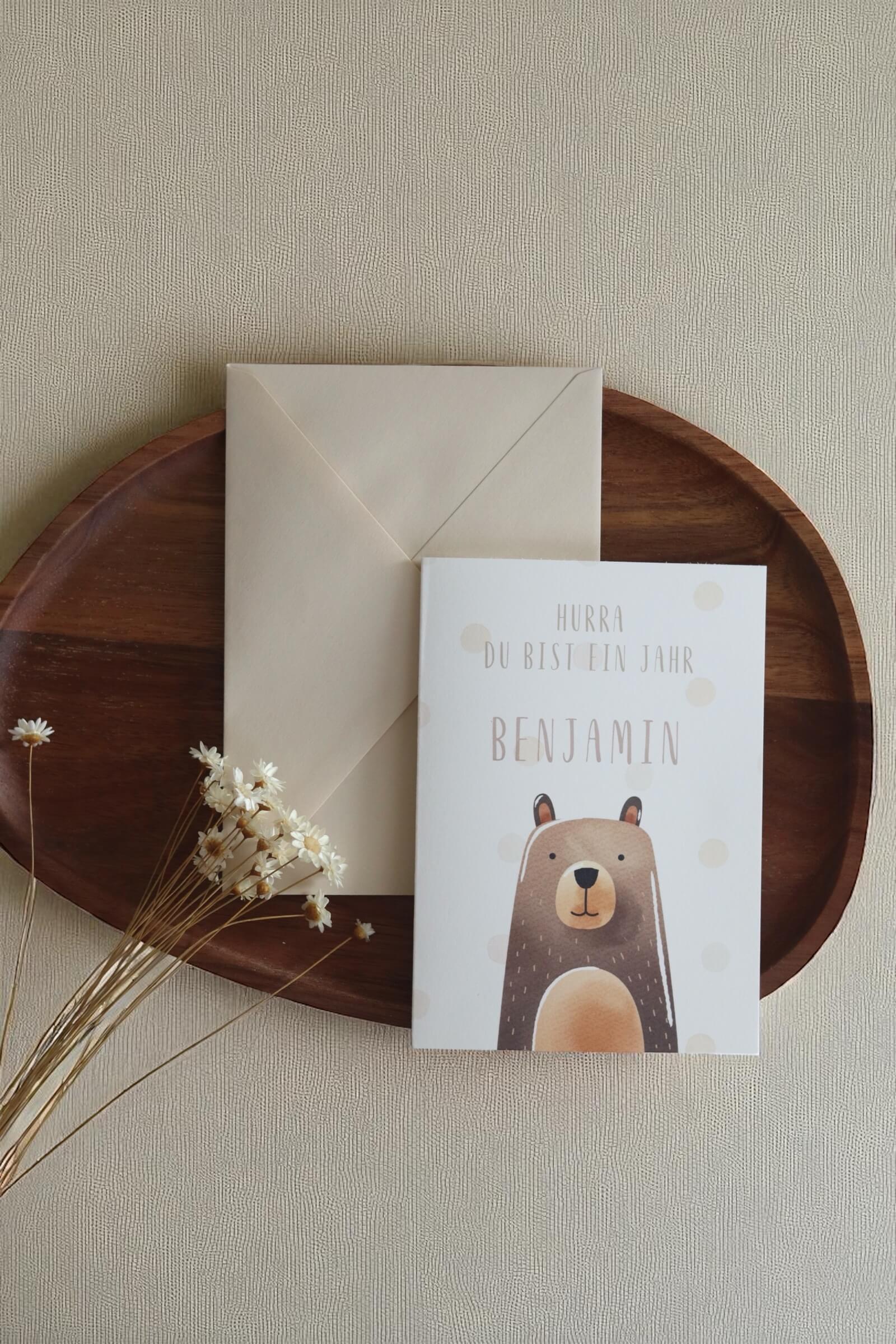 Grußkarte im Postkarten format mit Bärenmotiv und Namen daneben einer beiger Umschlag auf einem Holztablett und Trockenblumen