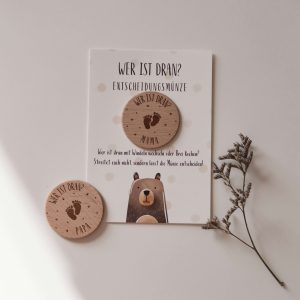 Karte mit einer Entscheidungsmünze aus Holz mit der Gravur "wer ist dran? Mama" vor neutralem Hintergrund