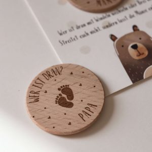 Karte mit einer Entscheidungsmünze aus Holz mit der Gravur "wer ist dran? Mama" vor neutralem Hintergrund
