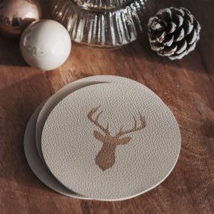 Beige runde Untersetzer aus Kunstleder mit einem Rentiermotiv auf einem dunklen Holzuntergrund und weihnachtlicher Dekoobjekte
