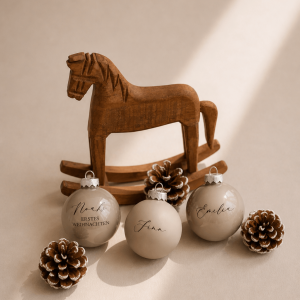 Drei beige Christbaumkugeln mit Namen graviert in einem minimalistischem Hintergrund mit Tannenzapfen und einem Holzschaukelpferd