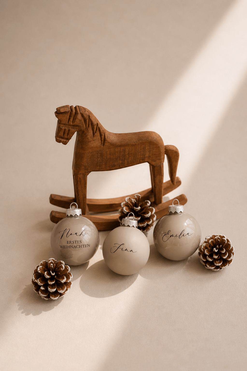 Drei beige Christbaumkugeln mit Namen graviert in einem minimalistischem Hintergrund mit Tannenzapfen und einem Holzschaukelpferd