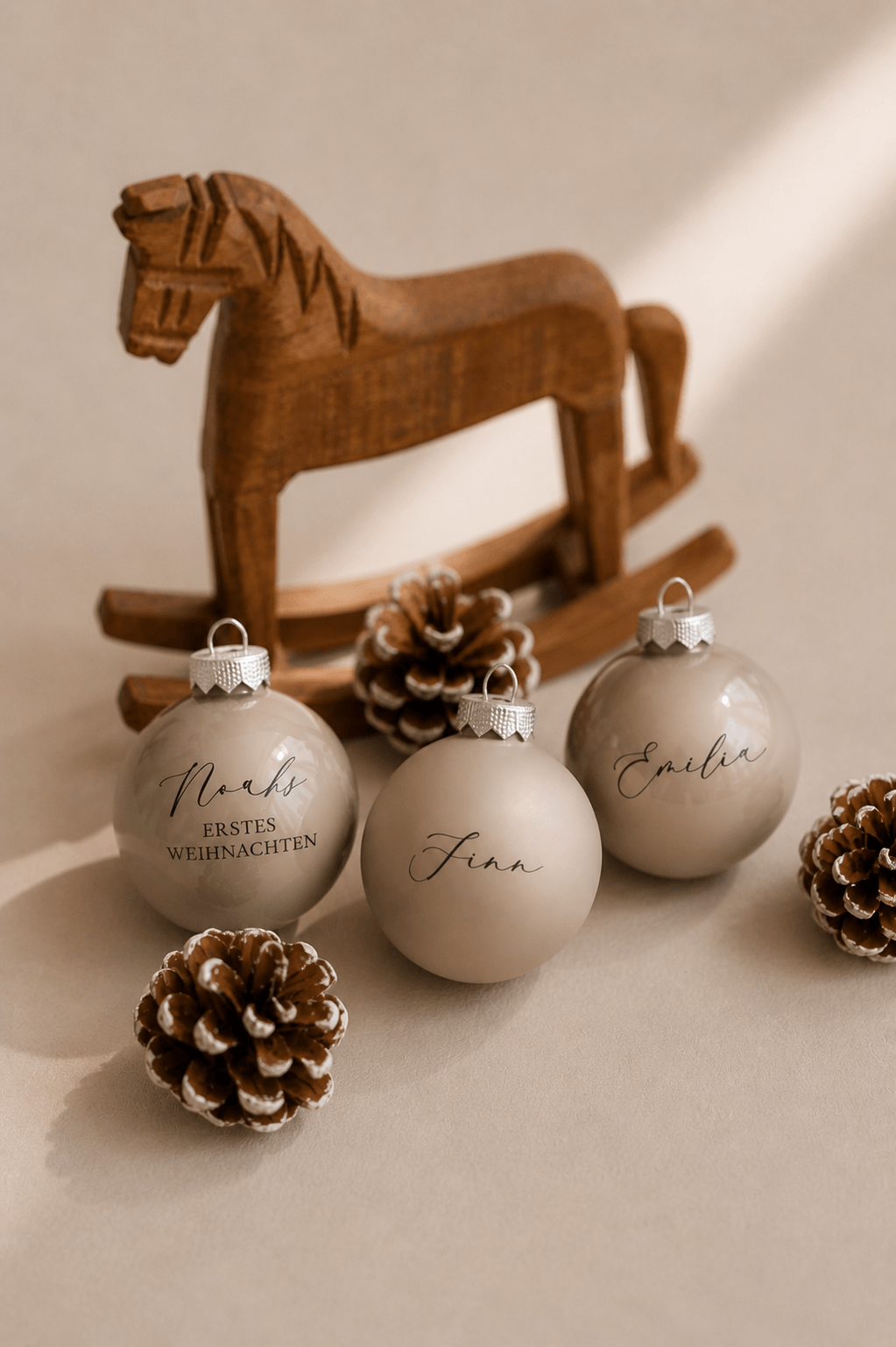Drei beige Christbaumkugeln mit Namen graviert in einem minimalistischem Hintergrund mit Tannenzapfen und einem Holzschaukelpferd