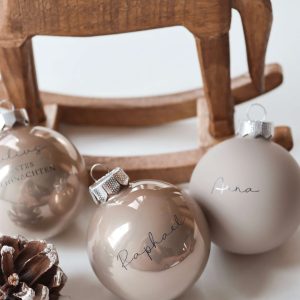 drei beigefarbene Christbaumkugeln aus Glas mit Namensgravur dahinter eine Schaukelpferd aus dunklem Holz und einem Tannenzapfen in minimalistischem Hintergrund