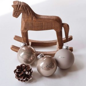 drei beigefarbene Christbaumkugeln aus Glas mit Namensgravur dahinter eine Schaukelpferd aus dunklem Holz und einem Tannenzapfen in minimalistischem Hintergrund