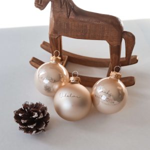 drei champagnerfarbene Christbaumkugeln aus Glas mit Namensgravur dahinter eine Schaukelpferd aus dunklem Holz und einem Tannenzapfen in minimalistischem Hintergrund