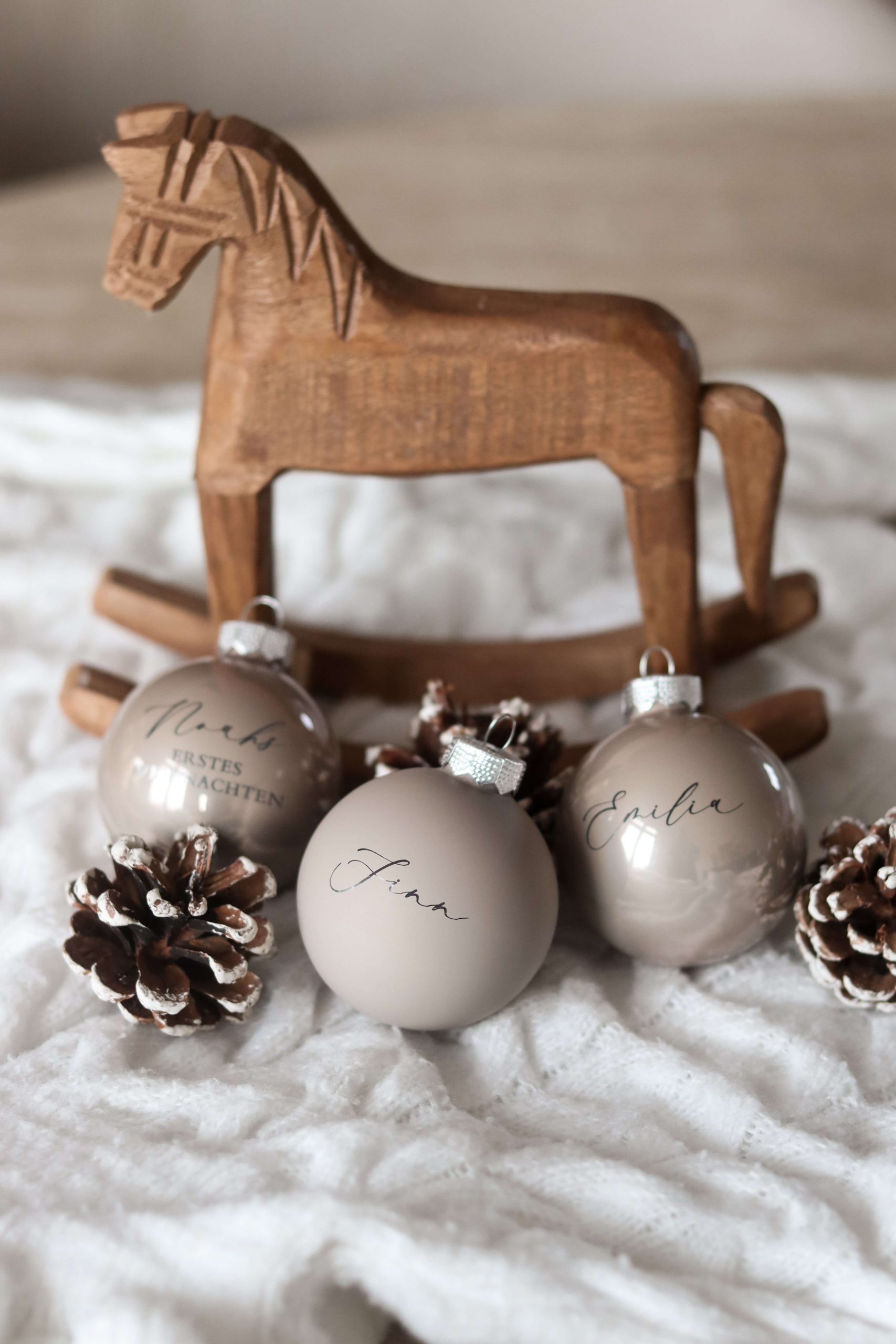 Beige Christbaumkugeln mit Namensgravur auf einem weißen Stoffuntergrund mit Tannenzapfen als Deko und einem Holzschaukelpferd im Hintergrund