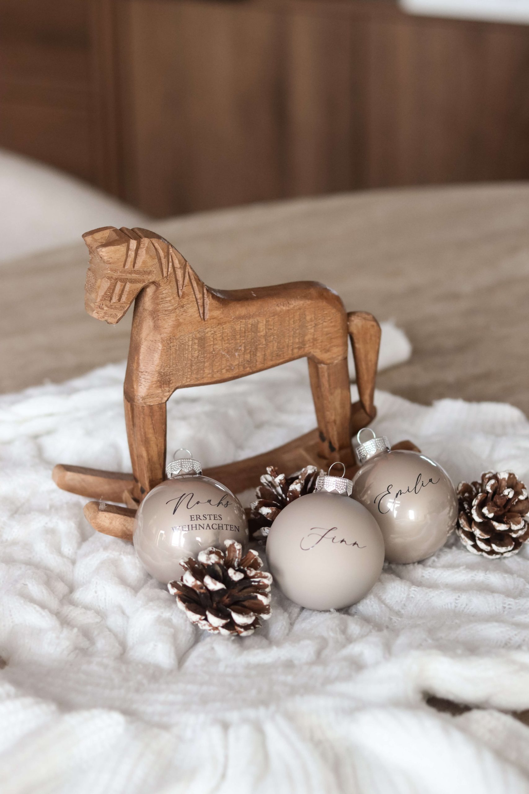 Beige Christbaumkugeln mit Namensgravur auf einem weißen Stoffuntergrund mit Tannenzapfen als Deko und einem Holzschaukelpferd im Hintergrund