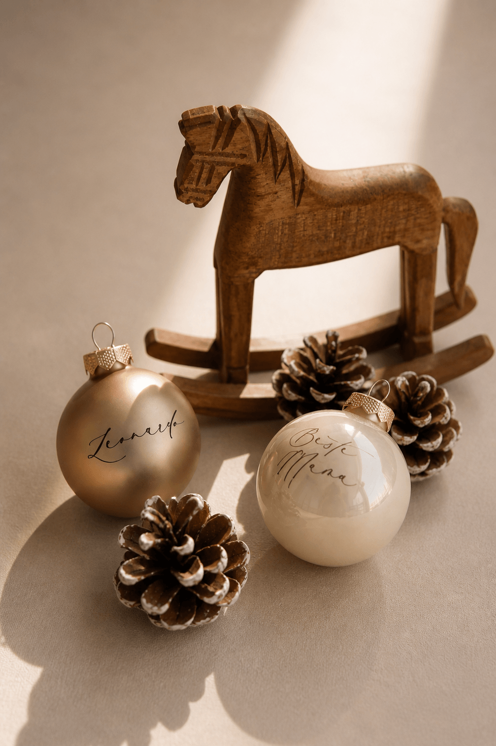 Zwei champagnerfarbene Christbaumkugeln mit Namen graviert in einem minimalistischem Hintergrund mit Tannenzapfen und einem Holzschaukelpferd