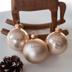 drei champagnerfarbene Christbaumkugeln aus Glas mit Namensgravur dahinter eine Schaukelpferd aus dunklem Holz und einem Tannenzapfen in minimalistischem Hintergrund