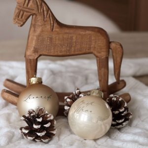 Champagnerfarbene Christbaumkugeln mit Namensgravur auf einem weißen Stoffuntergrund mit Tannenzapfen als Deko und einem Holzschaukelpferd im Hintergrund