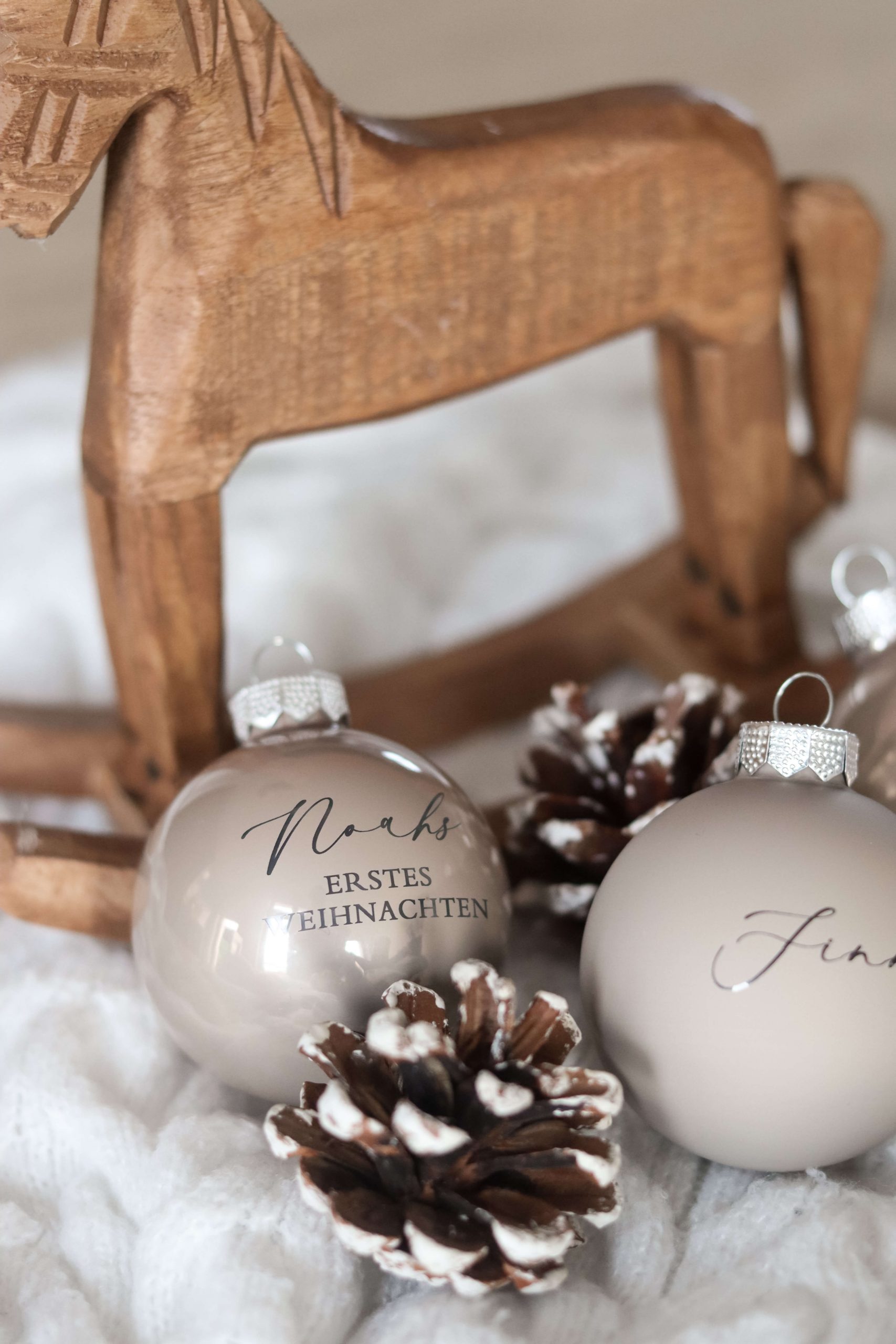 Beige Christbaumkugeln mit Namensgravur auf einem weißen Stoffuntergrund mit Tannenzapfen als Deko und einem Holzschaukelpferd im Hintergrund