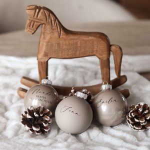 Beige Christbaumkugeln mit Namensgravur auf einem weißen Stoffuntergrund mit Tannenzapfen als Deko und einem Holzschaukelpferd im Hintergrund