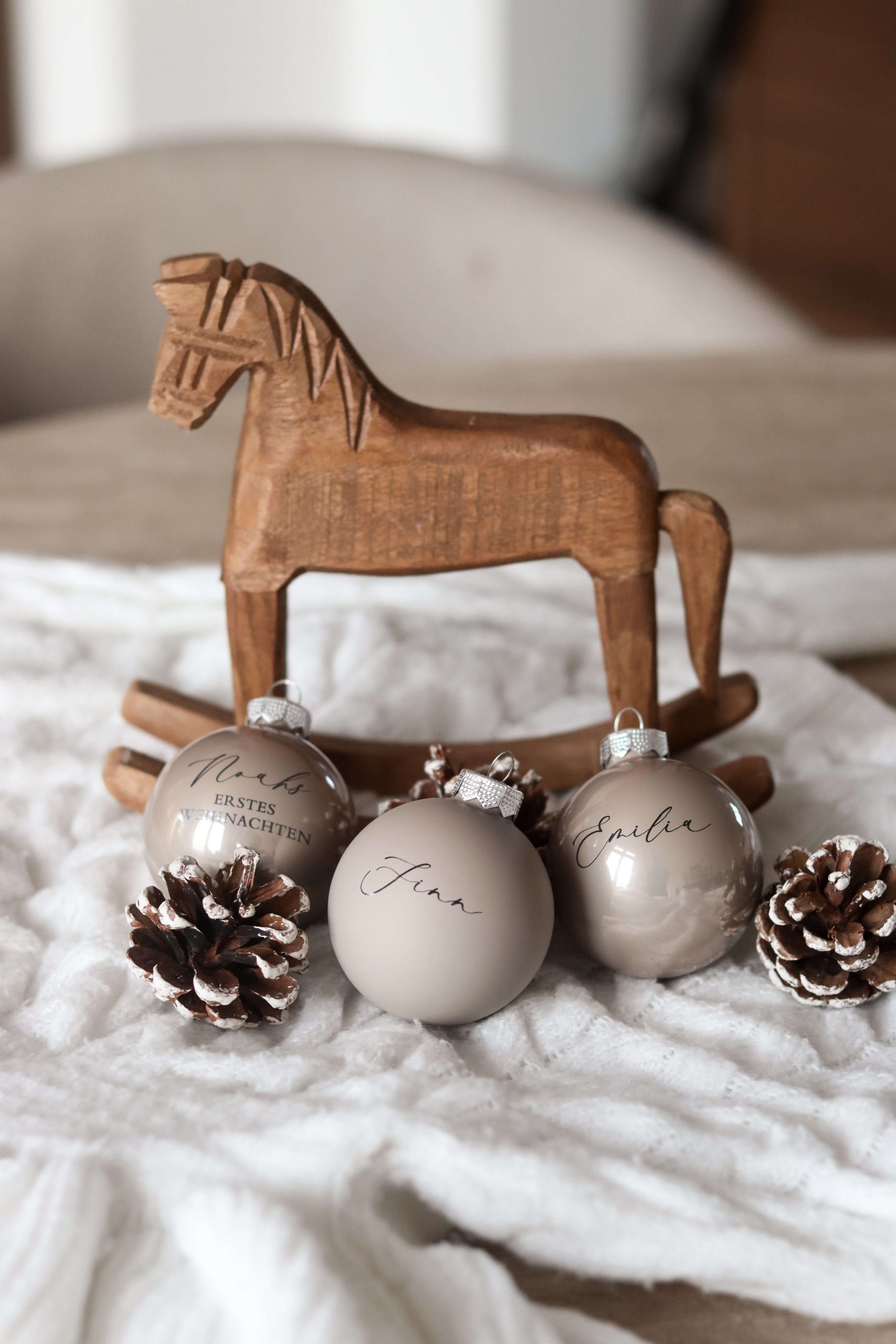 Beige Christbaumkugeln mit Namensgravur auf einem weißen Stoffuntergrund mit Tannenzapfen als Deko und einem Holzschaukelpferd im Hintergrund