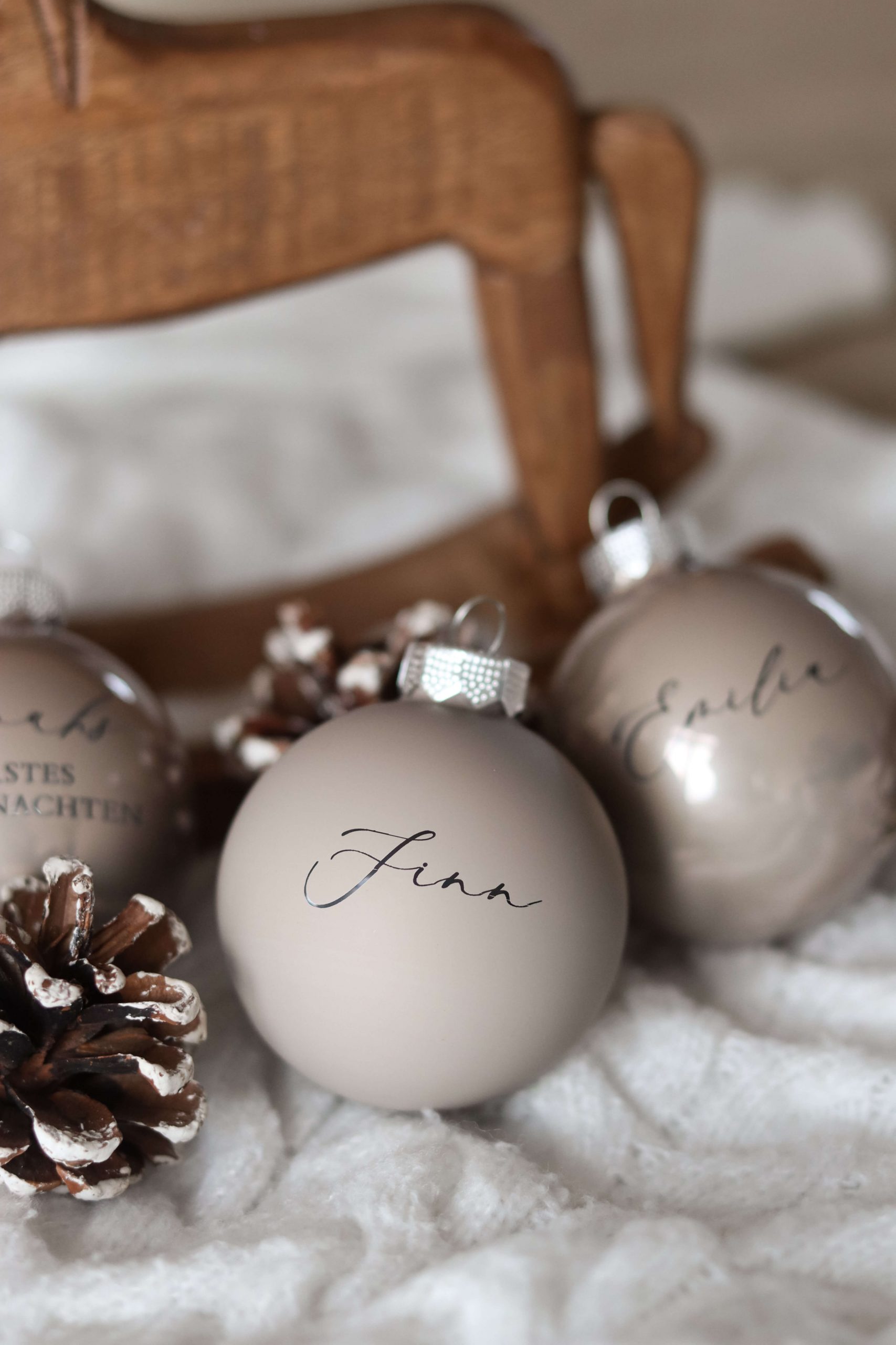 Beige Christbaumkugeln mit Namensgravur auf einem weißen Stoffuntergrund mit Tannenzapfen als Deko und einem Holzschaukelpferd im Hintergrund