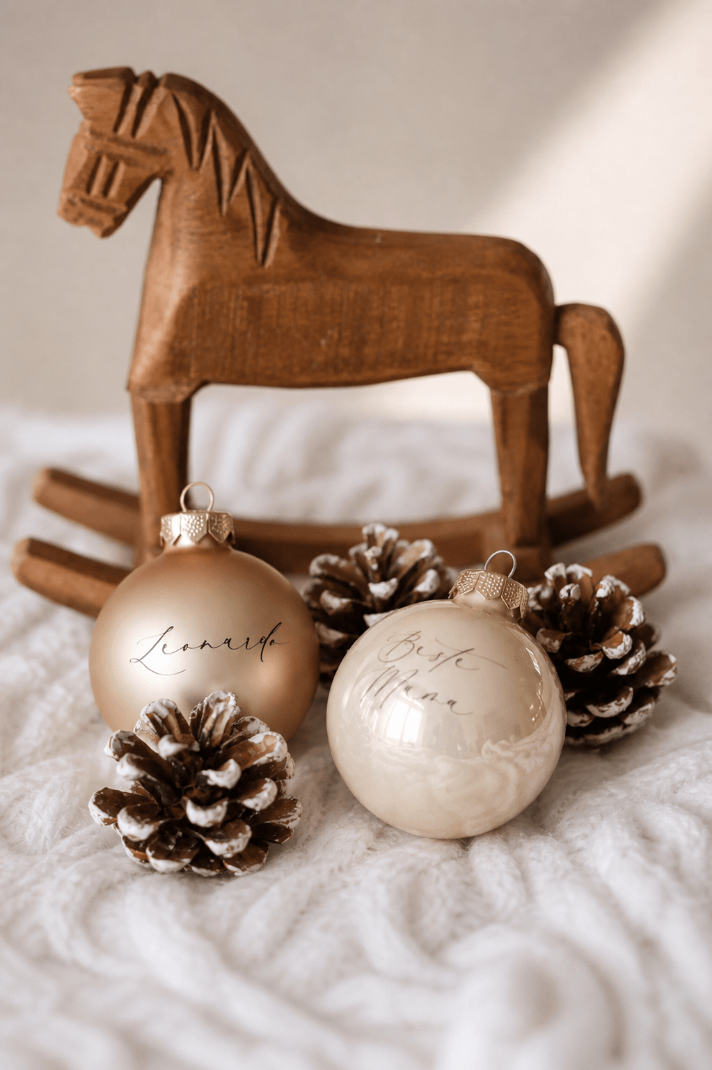 Zwei champagnerfarbene Christbaumkugeln mit Namen graviert in einem minimalistischem Hintergrund mit Tannenzapfen und einem Holzschaukelpferd
