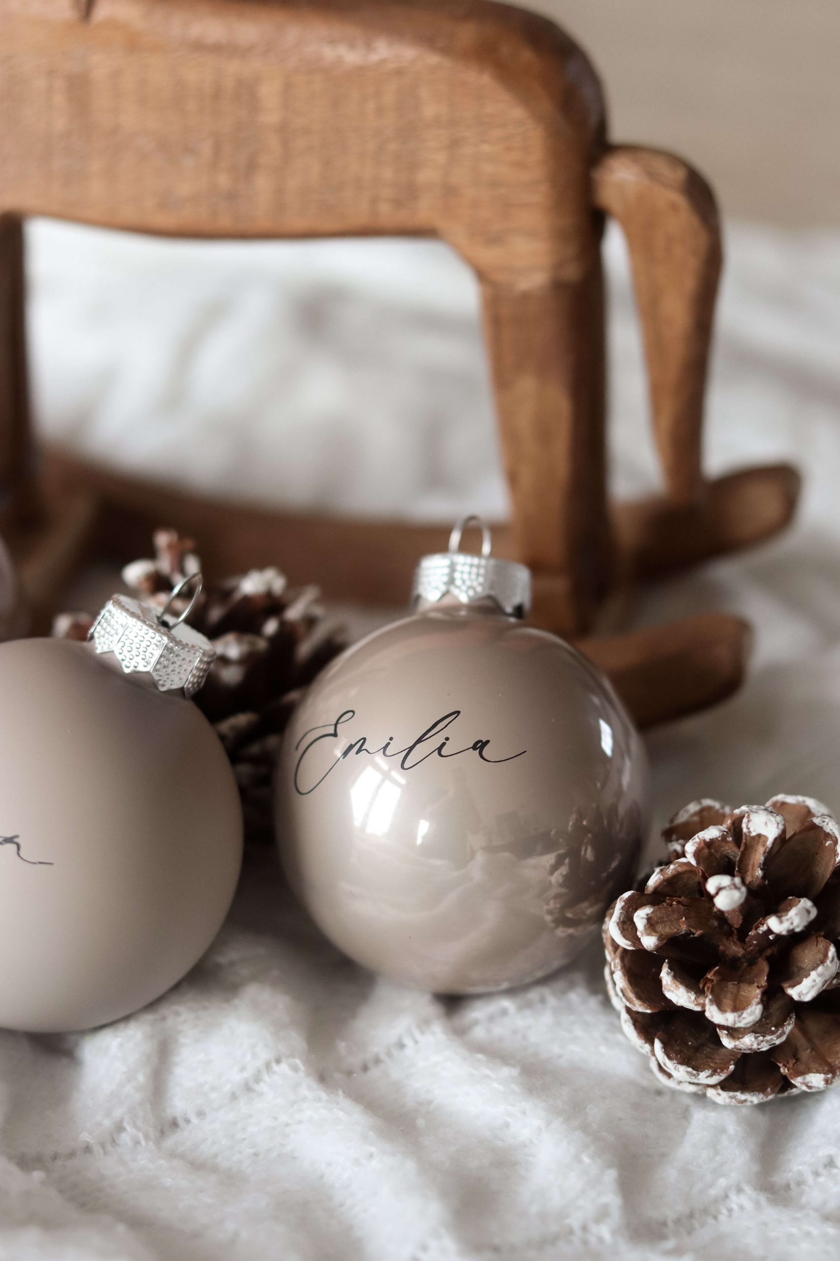 Beige Christbaumkugeln mit Namensgravur auf einem weißen Stoffuntergrund mit Tannenzapfen als Deko und einem Holzschaukelpferd im Hintergrund