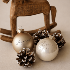 Zwei champagnerfarbene Christbaumkugeln mit Namen graviert in einem minimalistischem Hintergrund mit Tannenzapfen und einem Holzschaukelpferd