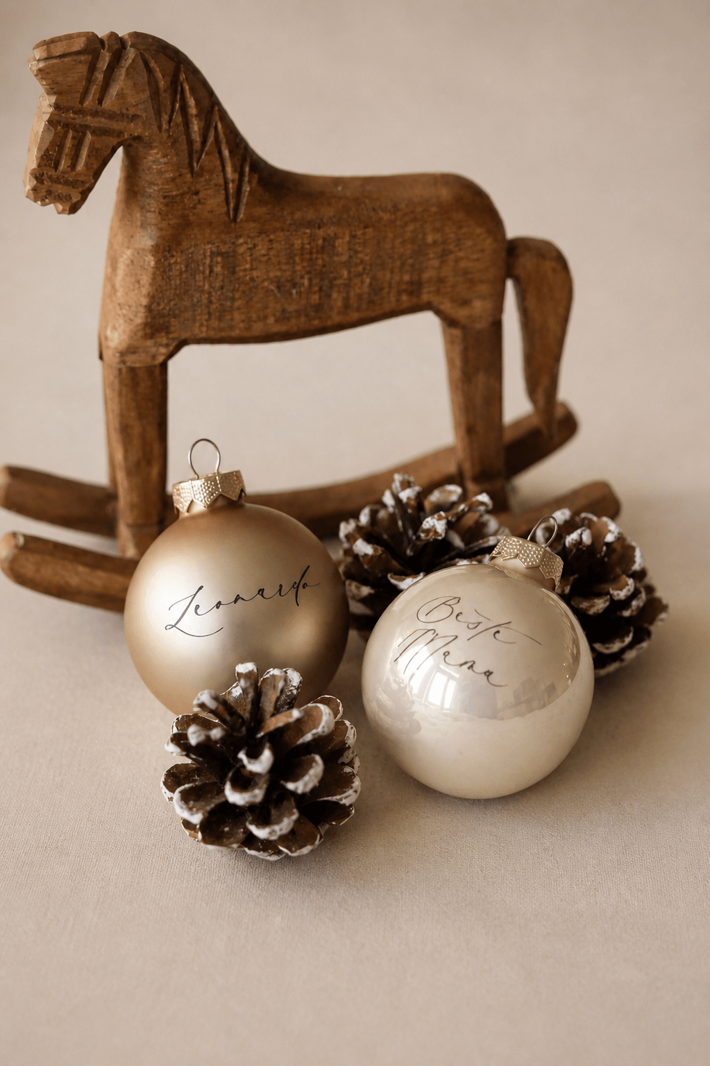 Zwei champagnerfarbene Christbaumkugeln mit Namen graviert in einem minimalistischem Hintergrund mit Tannenzapfen und einem Holzschaukelpferd