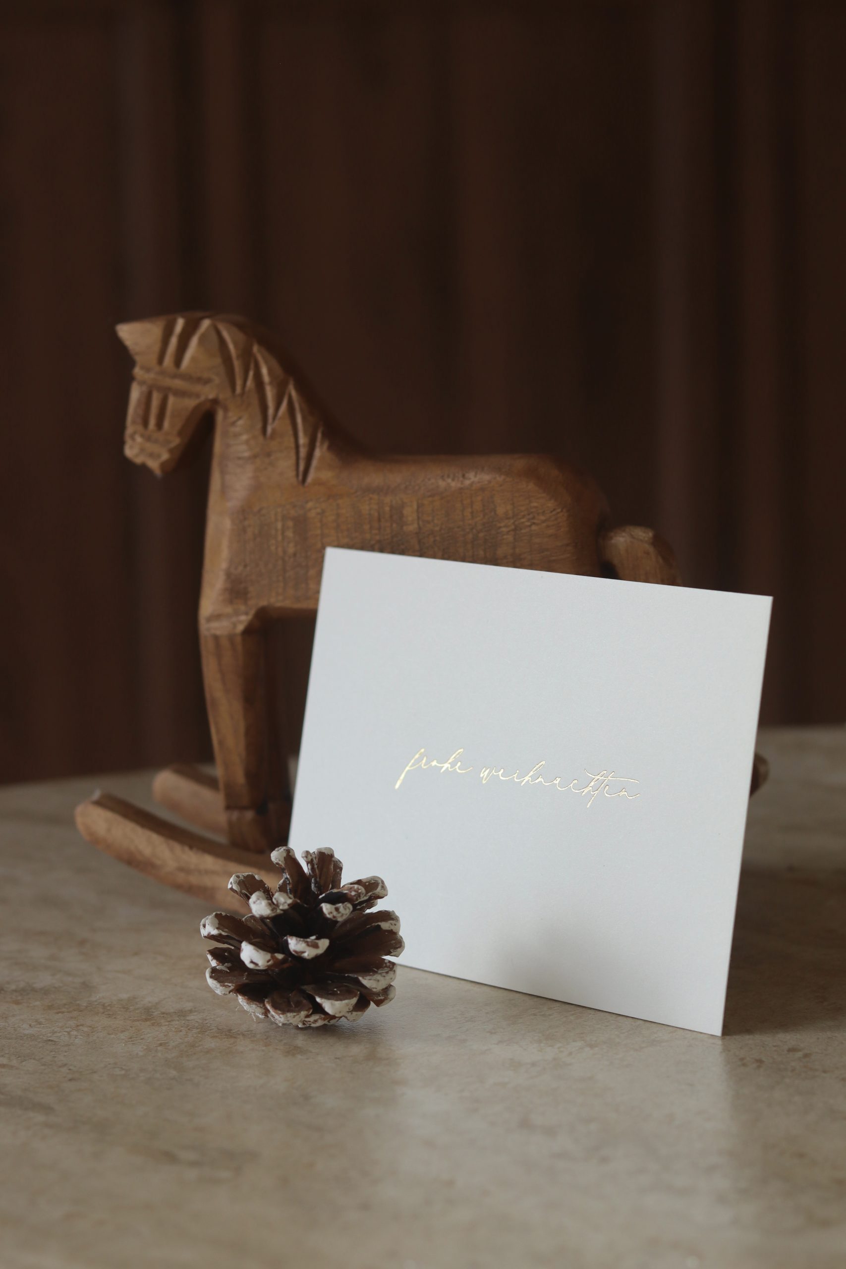 Weihnachtskarte im Querformat mit goldener Heißfolienprägung „Frohe Weihnachten“, dekoriert mit einem kleinen Holzschaukelpferd im Hintergrund.