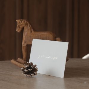 Weihnachtskarte im Querformat mit goldener Heißfolienprägung „Frohe Weihnachten“, dekoriert mit einem kleinen Holzschaukelpferd im Hintergrund.