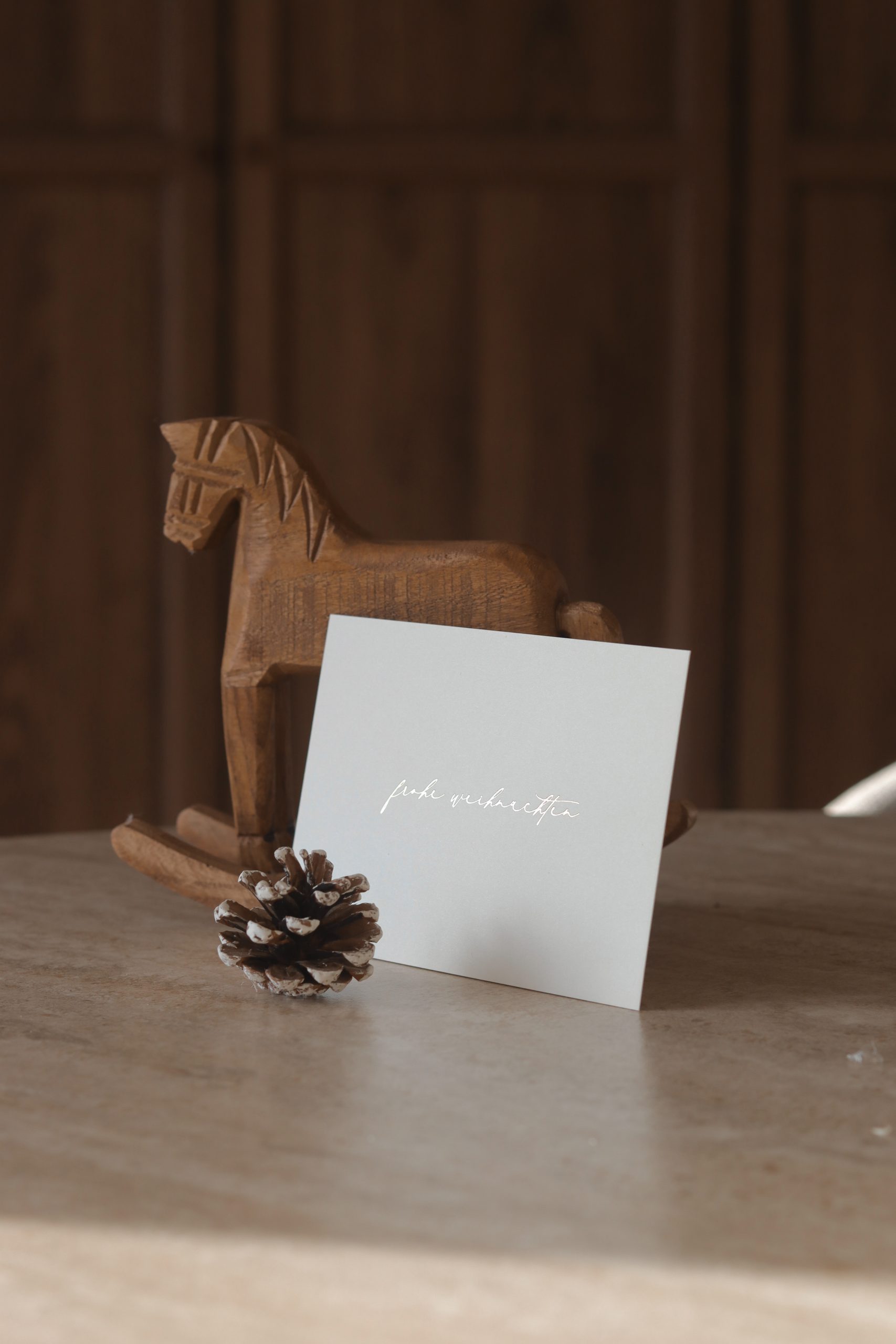 Weihnachtskarte im Querformat mit goldener Heißfolienprägung „Frohe Weihnachten“, dekoriert mit einem kleinen Holzschaukelpferd im Hintergrund.